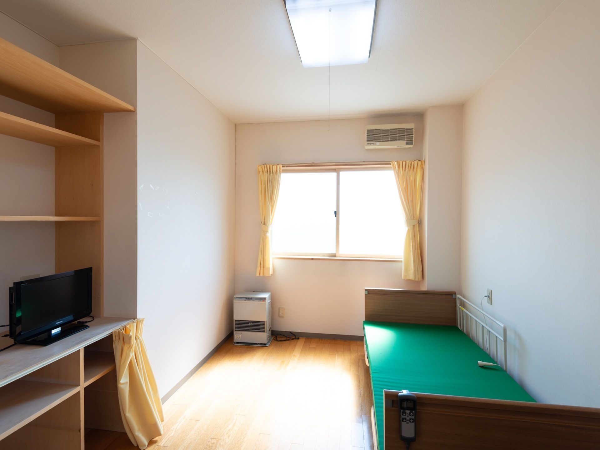 居室には窓があり、黄色いカーテンがかけられている。壁側にはベッドがある。ベッドの反対側の棚の上にはテレビが設置されている。