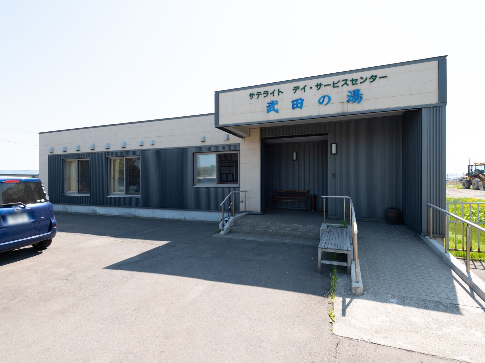 1階建ての建物になっていて、右側にはスロープや手すりが設置され、側には階段がある。建物の前は広くなっており、車が停められている。