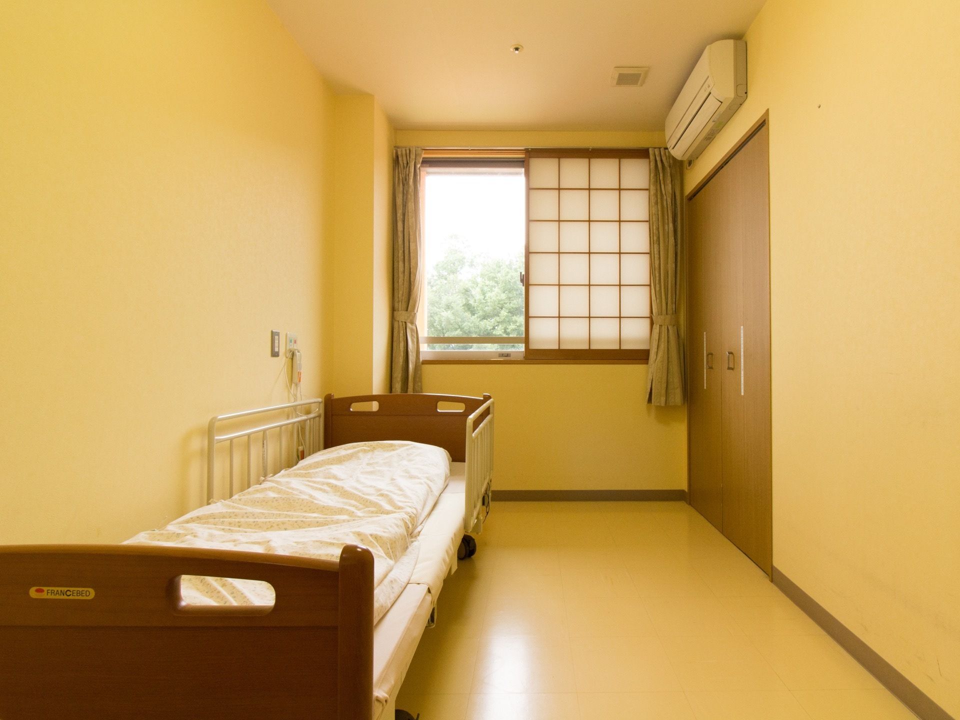 クローゼット、エアコン完備の１人用居室である。壁際に介護ベッドを置き、奥の窓には障子とカーテンの両方を取り付けている。