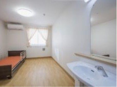 広々とした居室。手すりのついたベッドが置かれ、その上にはエアコンが設置されている。手前には洗面台もある。