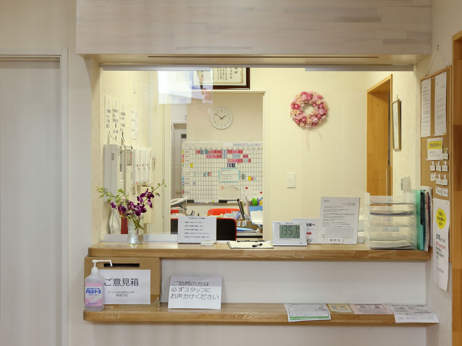 受付のカウンターは木目になっており、様々な書類やご意見箱、消毒用アルコール、時計やお花などが飾られている。