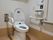温水洗浄機能付きのトイレである。壁にはナースコールやＬ字型の手すりが設置されており、上部には収納棚がある。