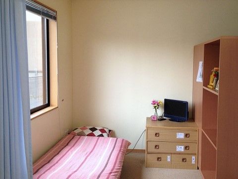 手前に薄い水色の仕切りカーテンがかかっている部屋で、窓はブラインドになっている。窓際のベッドの隣にはテレビが乗った小さなタンスが置いてある。