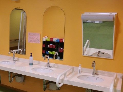 洗面台は３つあり、それぞれに液体の手洗い石けんが置かれている。中央の鏡にコップや洗面用具が仕舞われた棚が映っている。