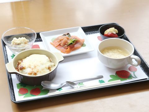 可愛らしいイチゴ柄のシートが敷かれたトレーには、ドリア、たまごスープ、メインの魚料理、小鉢などバラエティに富んだ料理が並べられている。