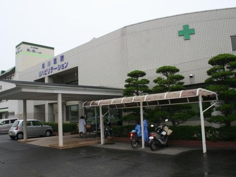 白い壁に緑の十字の模様と青字で書かれた医院の名称が表示されている。カーポートが設置され、バイクと自転車が停められている。