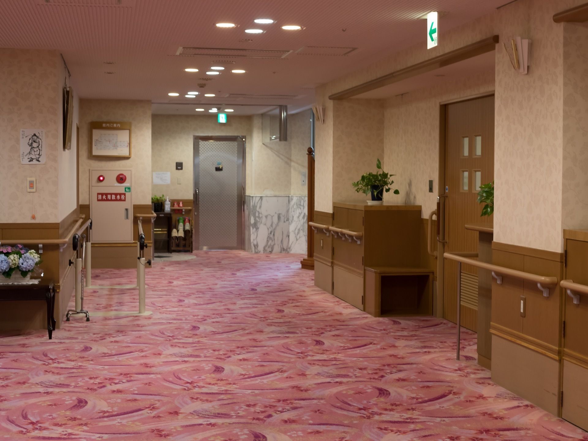 通路の壁には、手すりや防火設備が設置されている。床には華やかな印象のカーペットが敷かれ、歩行訓練用の平行バーが置かれている。
