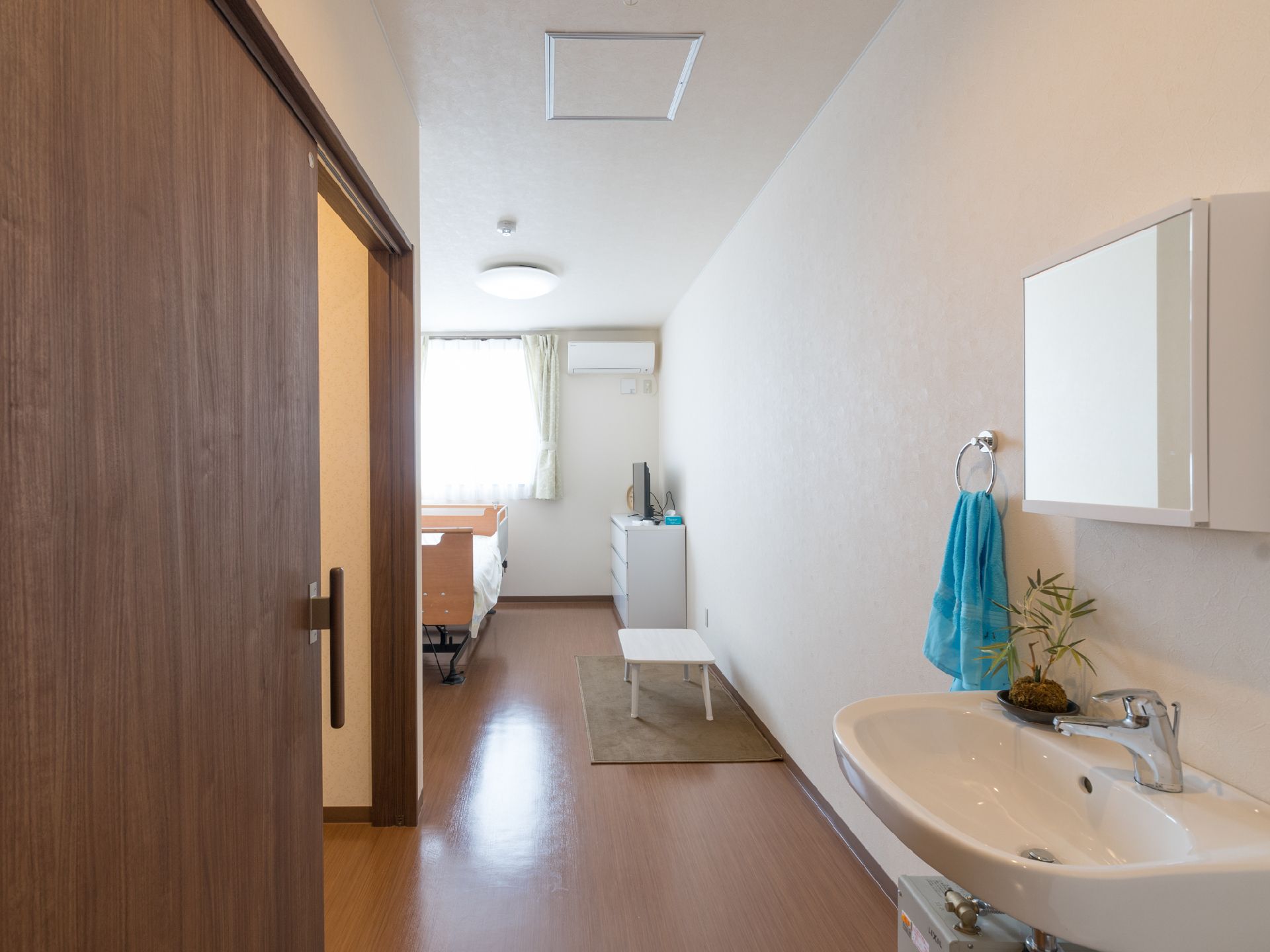 洗面台、テーブル、ベッド、タンスがある。冷暖房が完備されている。トイレドアが引き戸式である。窓がある。