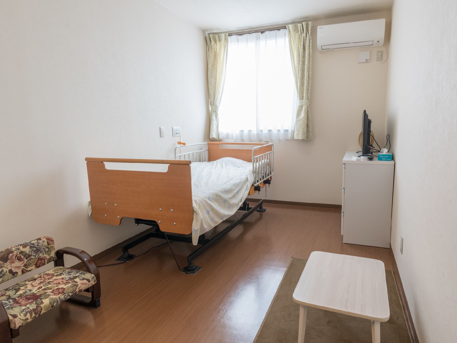 ベッド、座卓、椅子が配置されている。腰高窓があり、レースと柄物のカーテンが吊るされている。タンスの上にテレビが載せられている。