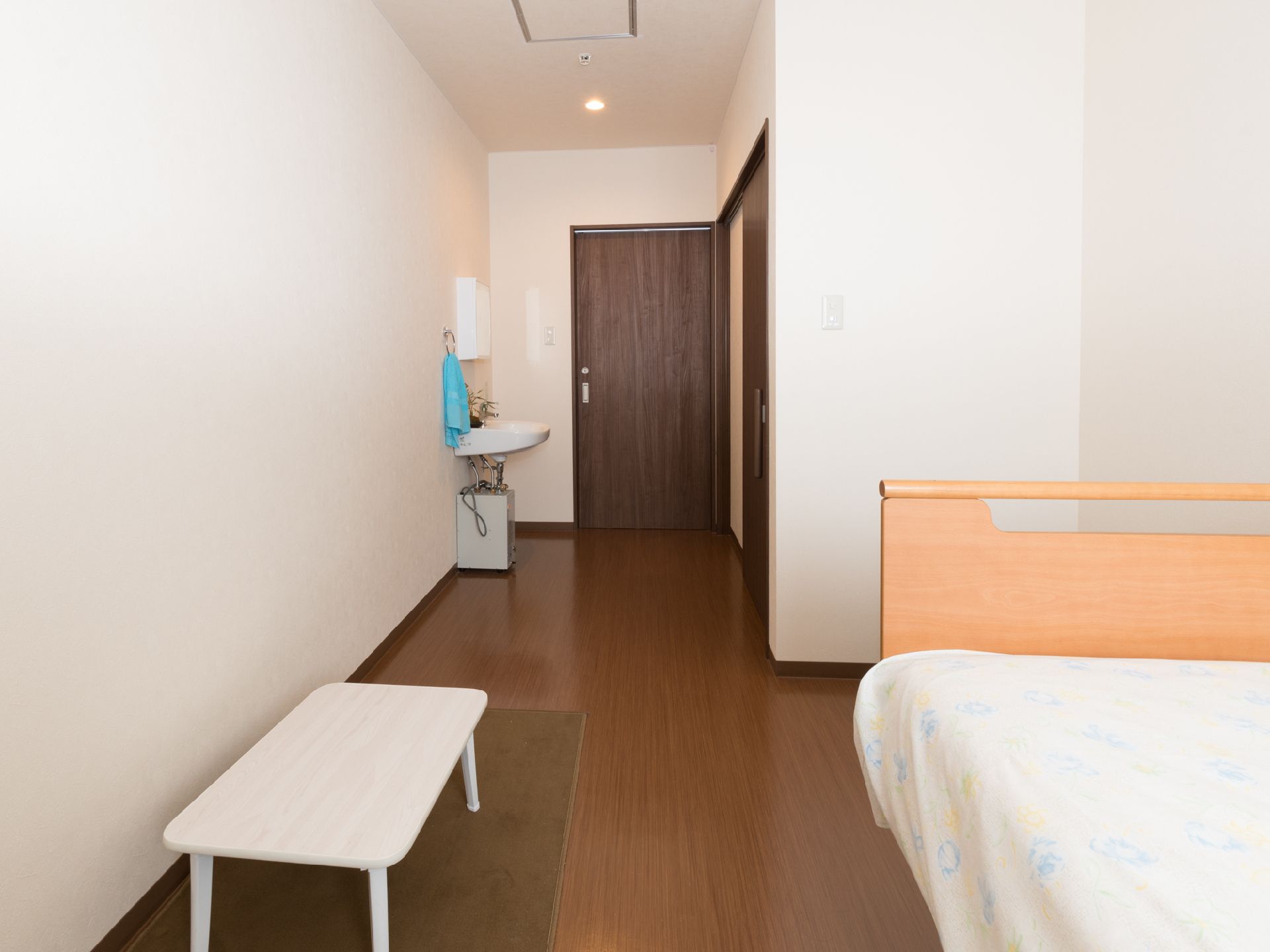 ベッド、洗面台、テーブルがある。入り口ドアとトイレのドアが引き戸式である。バリアフリー。壁は白く、床やドアは焦げ茶色の配色である。