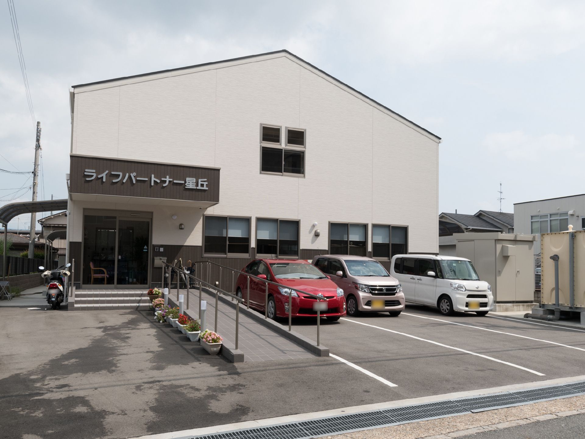スロープ横に花のプランターを置いている。建物隣に屋根付きの自転車、バイク置き場がある。入り口前に傘立てがある。
