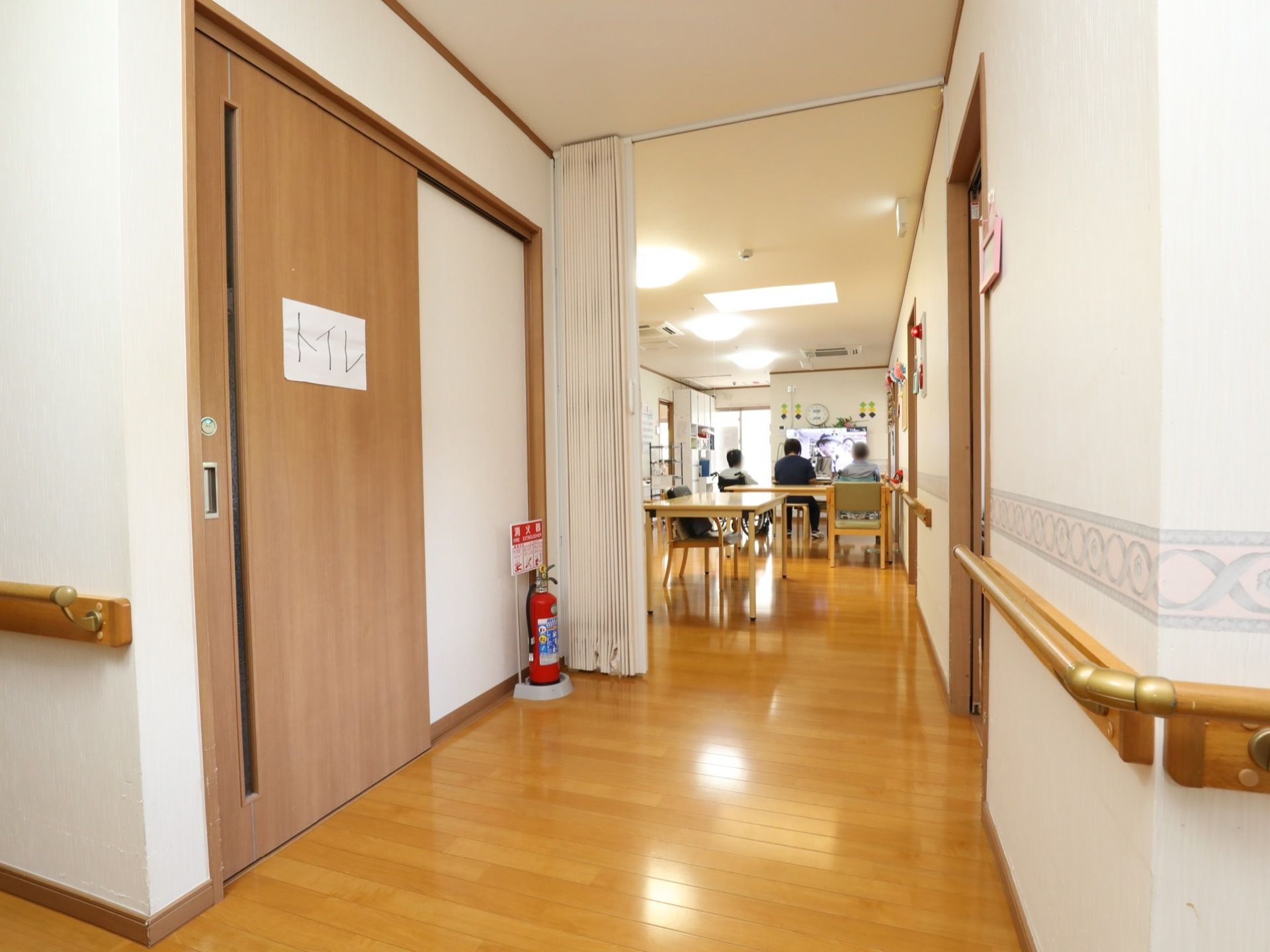 ライトブラウンの床の広い廊下がある。壁には手すりがあり、大きな扉のトイレがある。その先は食堂へと続いている。