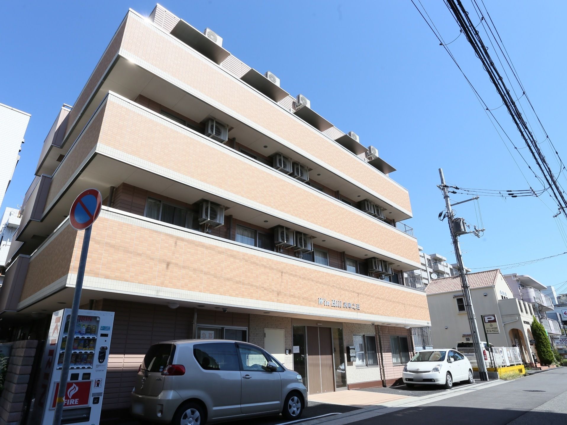 施設建物を外から見上げたところ。前の敷地には自動車が２台停められるスペースがあり、左には自動販売機が設置されている。