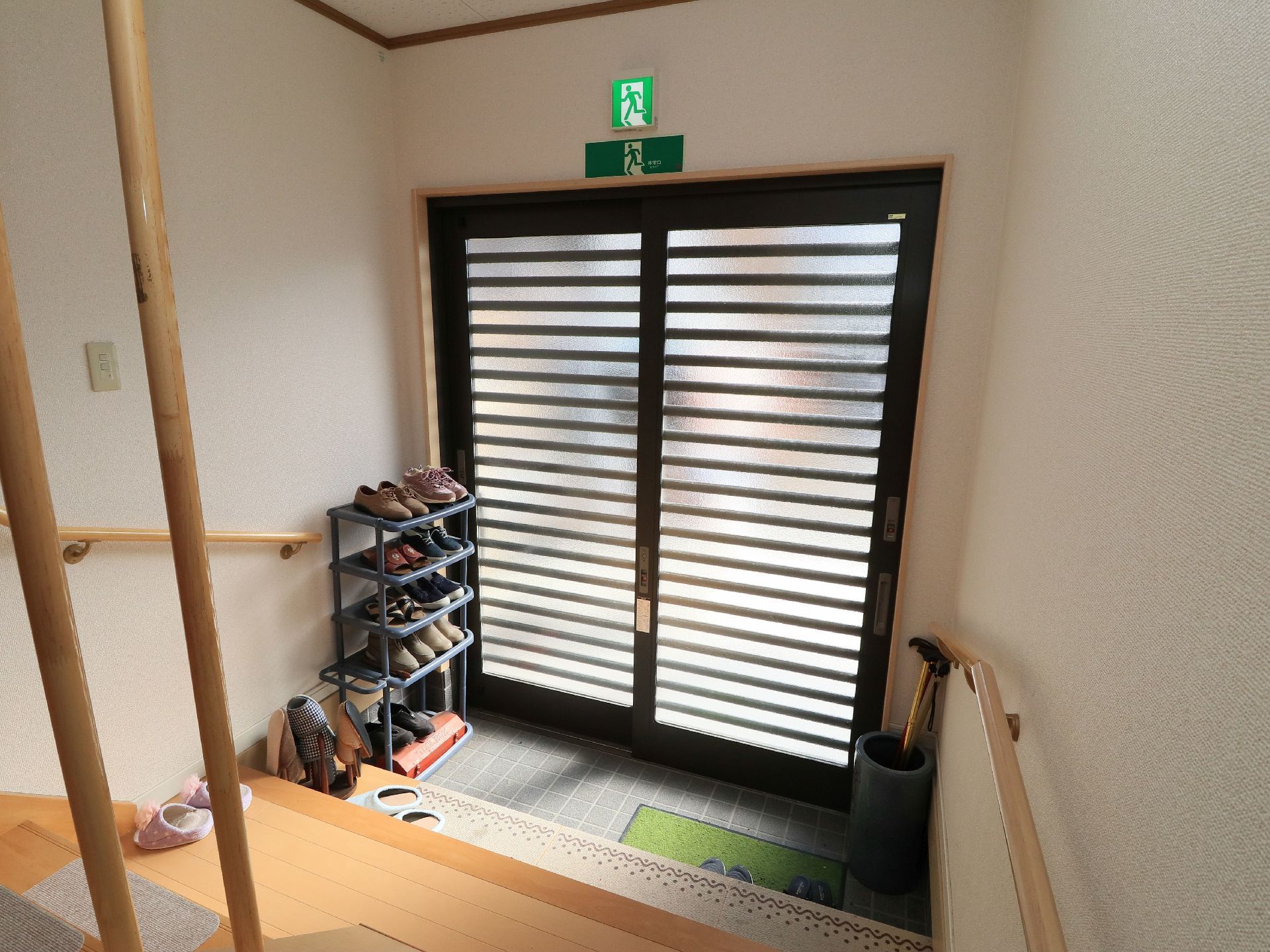 一般住宅のような雰囲気の玄関である。シューズラックや杖立てが壁際に置かれている。グリーンの玄関マットが敷かれている。