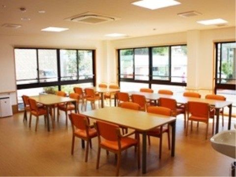 壁2面が大きな窓でとても開放感がある。テーブルにはオレンジ色の椅子が配置されている。手前の壁側にはシンプルな洗面台が設置されている。