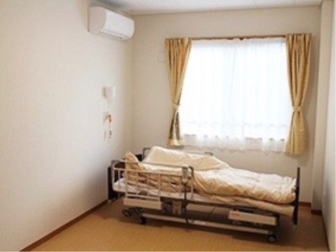 壁は清潔感のある白に近いアイボリー、床は落ち着いたベージュである。介護ベッドの頭側の壁にはナースコールがある。