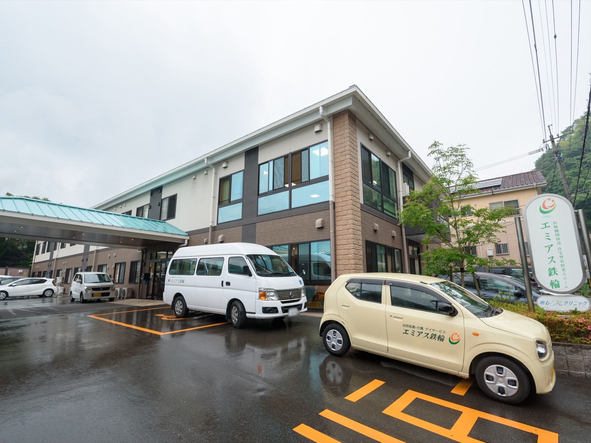 玄関は緑色の三角屋根となっている。建物前はアスファルトで舗装されており、施設専用車が複数台並んで停められている。