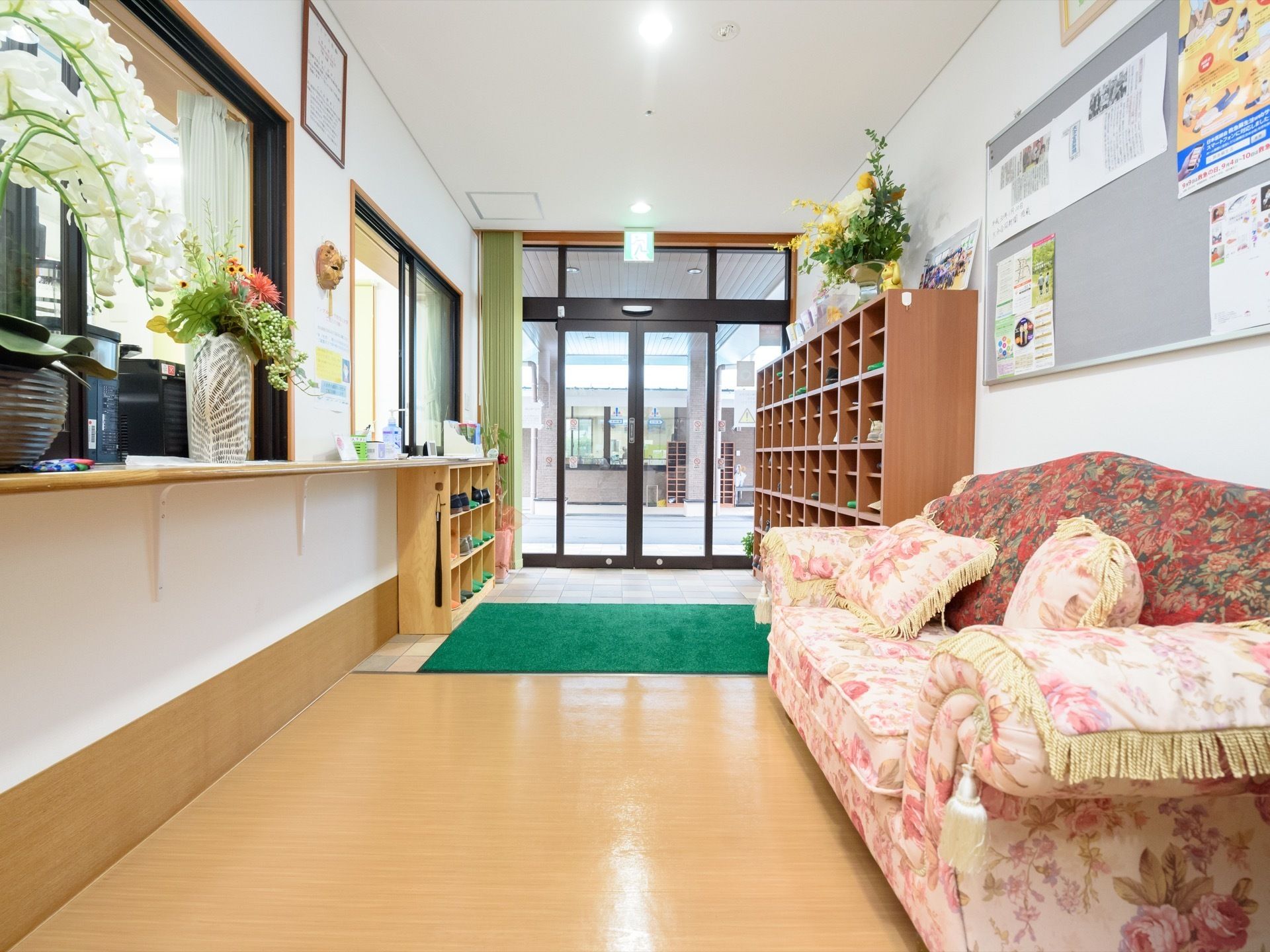 玄関横にある大きな靴箱の横には、花柄のソファーが置かれている。壁に掛けられた掲示板には、ポスターが複数貼られている。