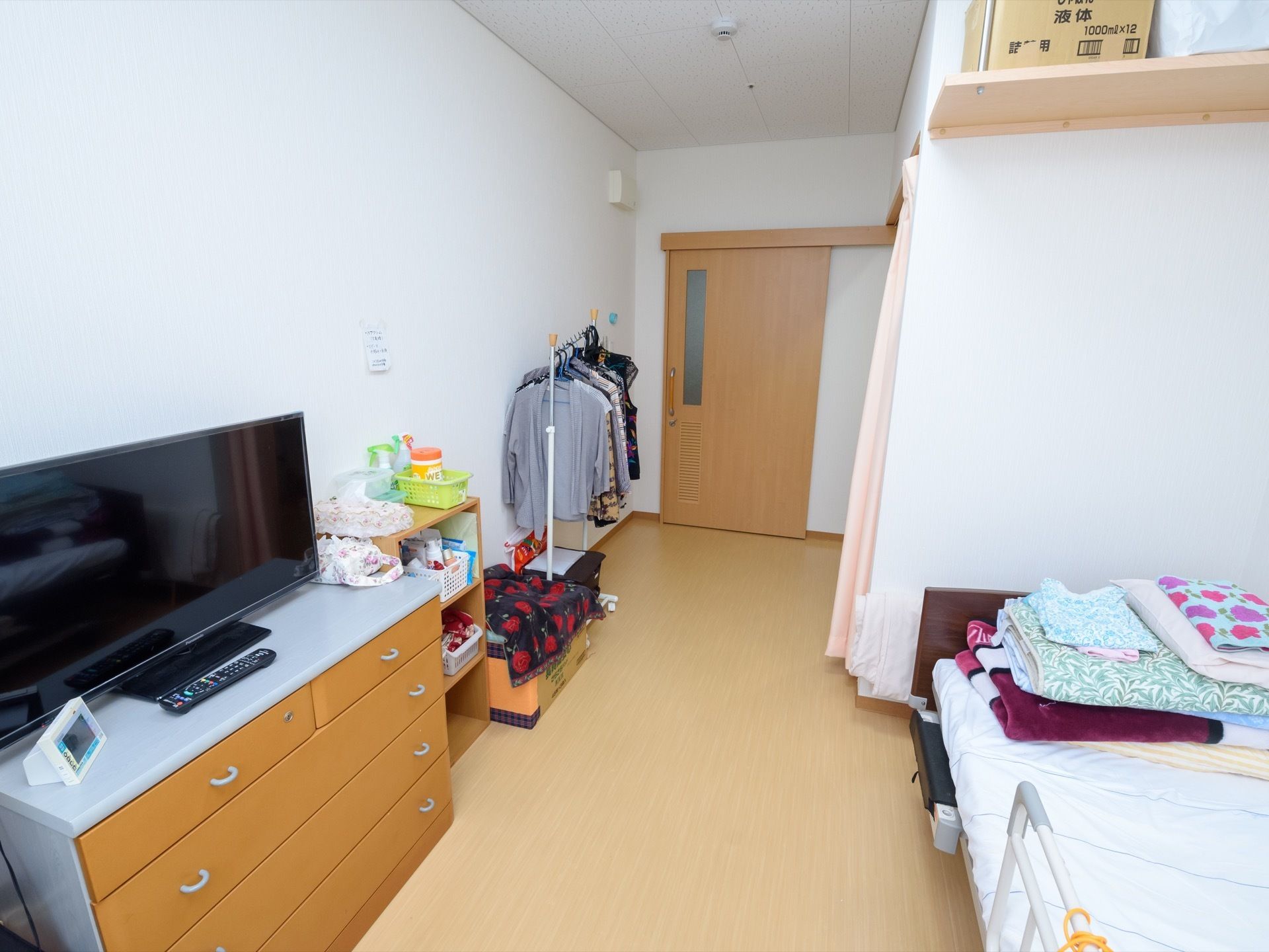 ベッドや洋服ダンス、テレビが設置されたワンルームの居室である。壁の上部には、棚が取り付けられている。
