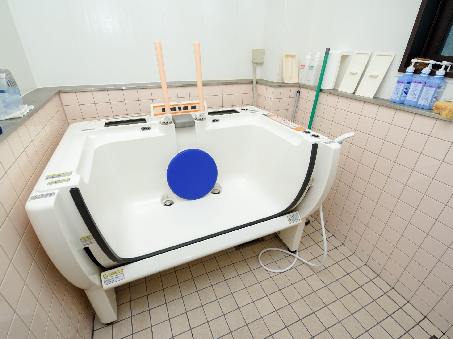 パネルの付いた白い機械浴層が置かれている。壁には窓とヒーターが取り付けられており、浴室周りの棚には消毒液などが置かれている。