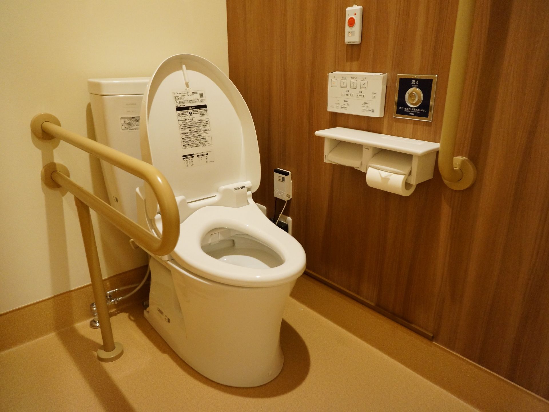 手すり付き洋式トイレ。緊急ブザーが壁に設置してある。予備トイレットペーパーホルダが設置してある。ウォッシュレットである。