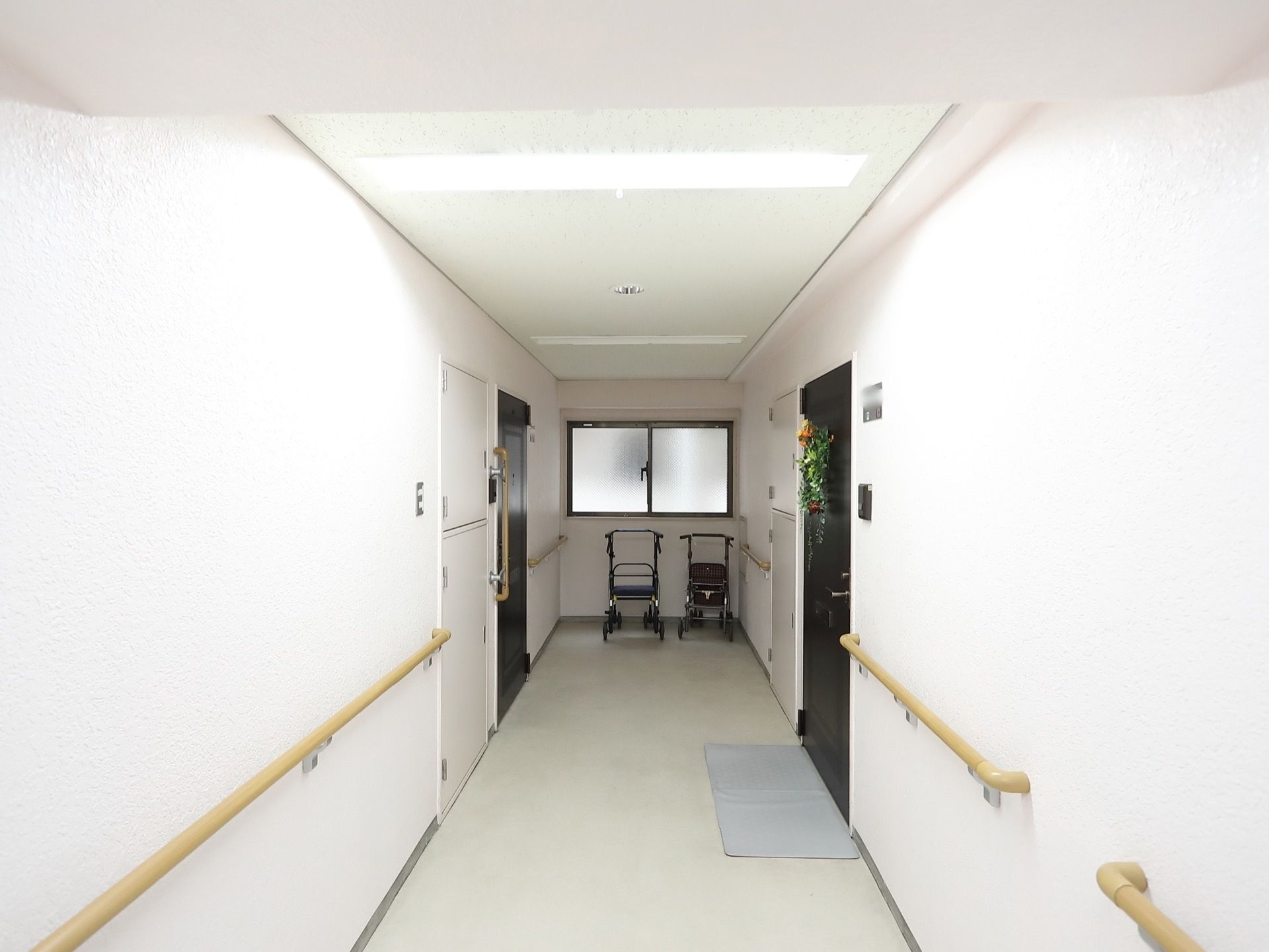 廊下は白色を基調とした明るい雰囲気になっている。居室の扉の前に灰色の足ふきマットが敷かれている。つきあたりに手押し車が置かれている。