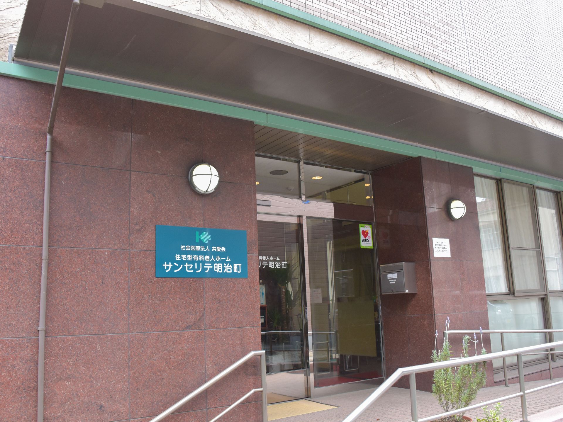 玄関は自動ドアで、入るまでにスロープが設置されている。壁には郵便ポストがあり、ガラスにはAEDのシールが貼られている。