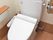 ウォシュレットのついた洋式トイレで、手を洗う場所が背部にある。手すりがついている。フローリングである。