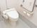 洋式トイレでウォシュレットがついたタイプである。手すりが設置してある。緊急ブザーが壁に設置してある。