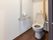 トイレの便器はきれいに磨かれており、床面は落ち着いた色合いのフローリングである。便器近くの壁には手すりやつかまり用の棒が備わっている。