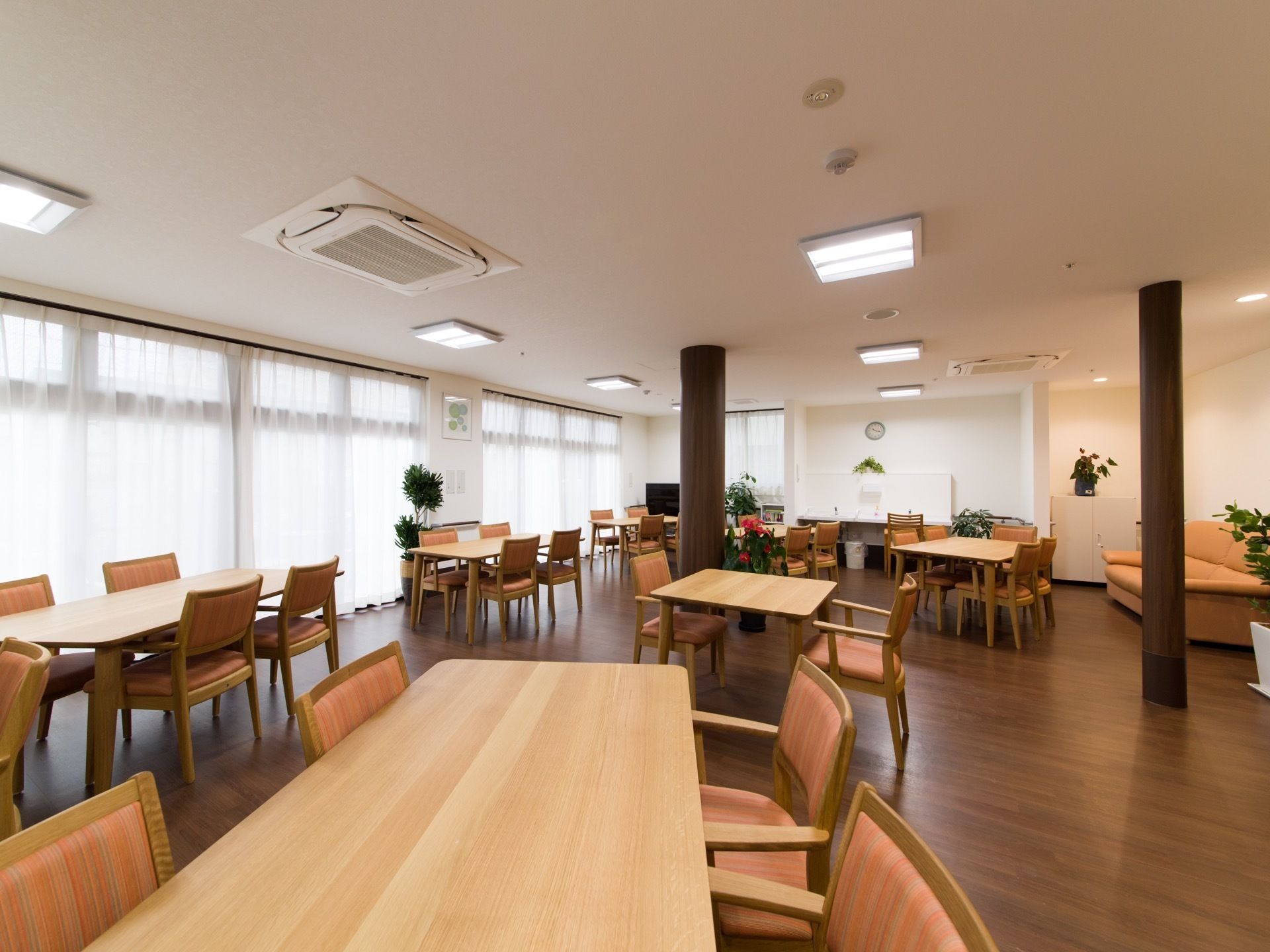 食堂の壁一面には大きな窓が並び、レースカーテンが引かれている。柱が2本あり、その奥に手洗い場とテレビがある。