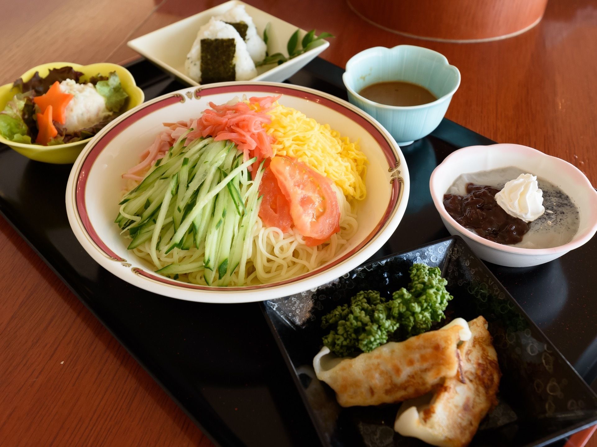 黒い四角い膳の上に、おにぎり２つ、サラダ、冷やし中華とたれ、揚げ餃子、デザートが並んでおり、どれも彩りや盛りつけが工夫されている。