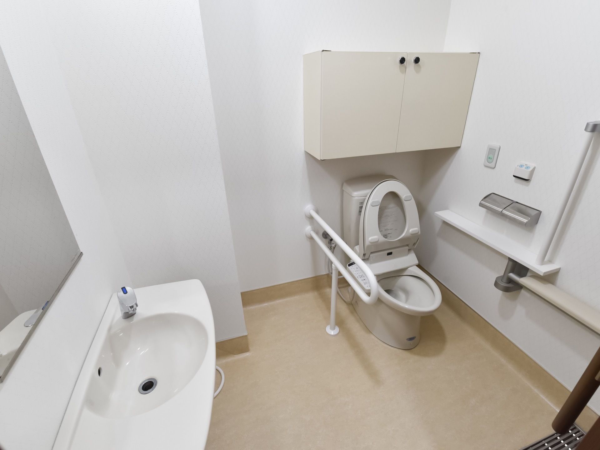 トイレ個室の様子。手元に操作ボタンが付いている、洋式トイレがある。壁に手すりがある。個室内に、手洗い場が併設されている。