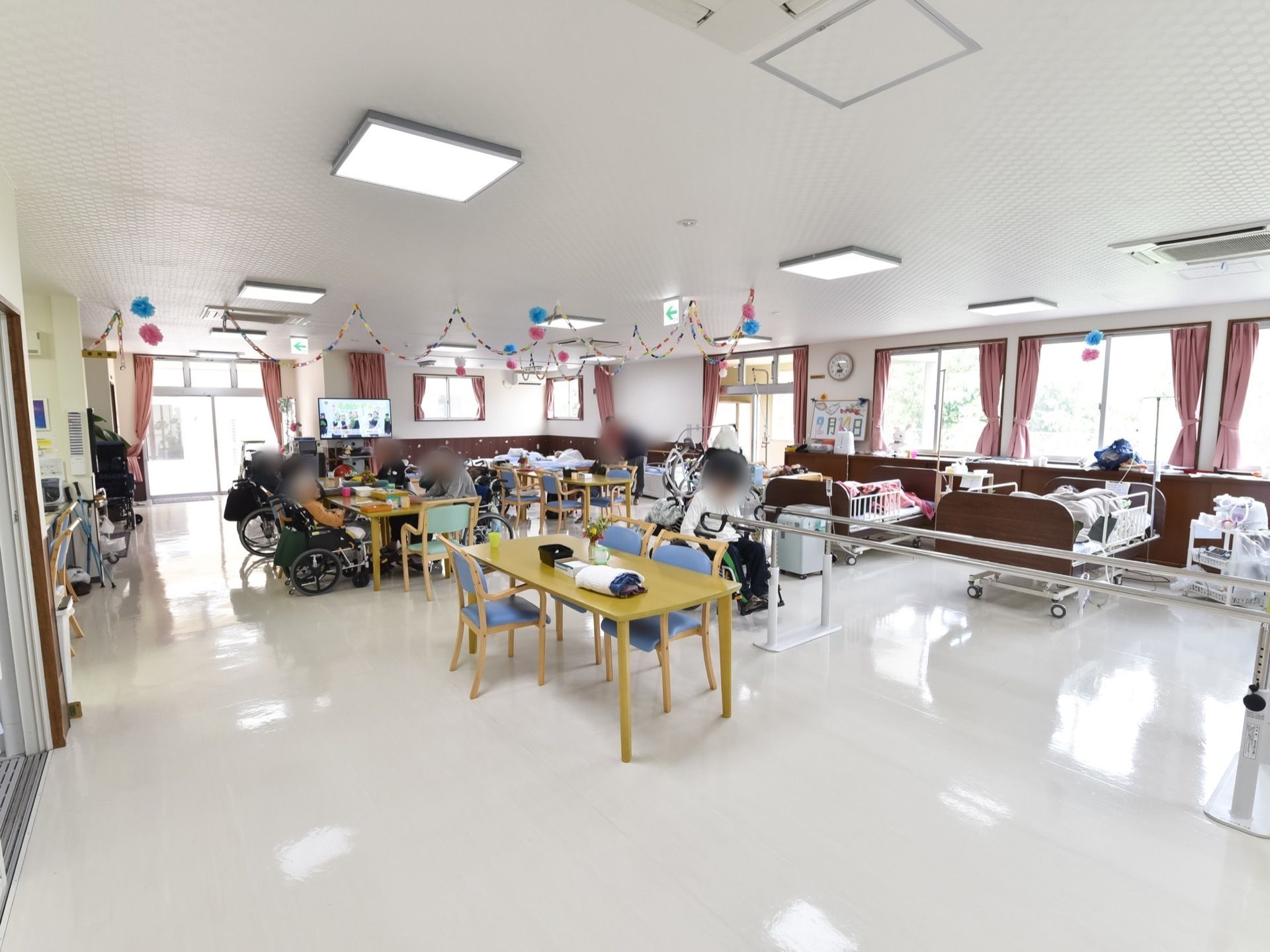 室内にテーブルが数台置かれ、入居者様たちが座っている。右手の窓際には、ベッドが2台置かれている。また、平行棒も設置されている。