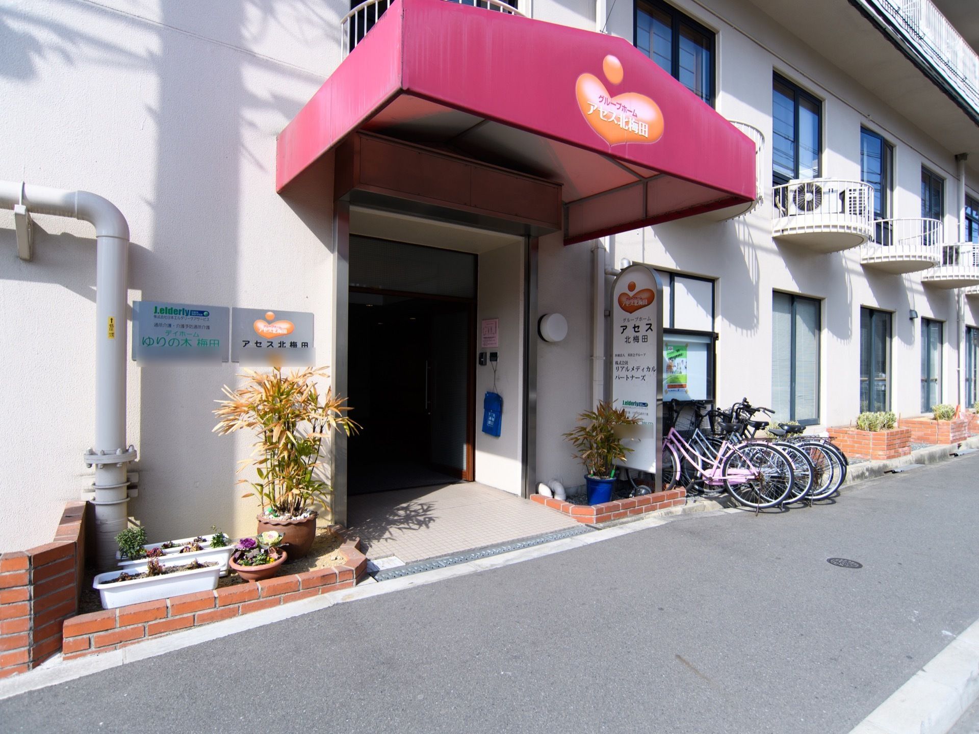 玄関の前方には、濃いピンク色のひさしが取り付けられている。その右端に看板があり、その横に自転車置き場がある。