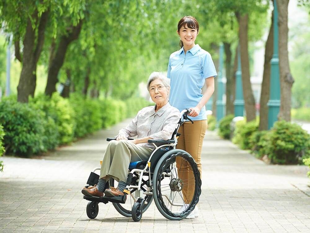 街路樹の下を散歩する車椅子に乗った高齢女性と介助をする女性ヘルパー