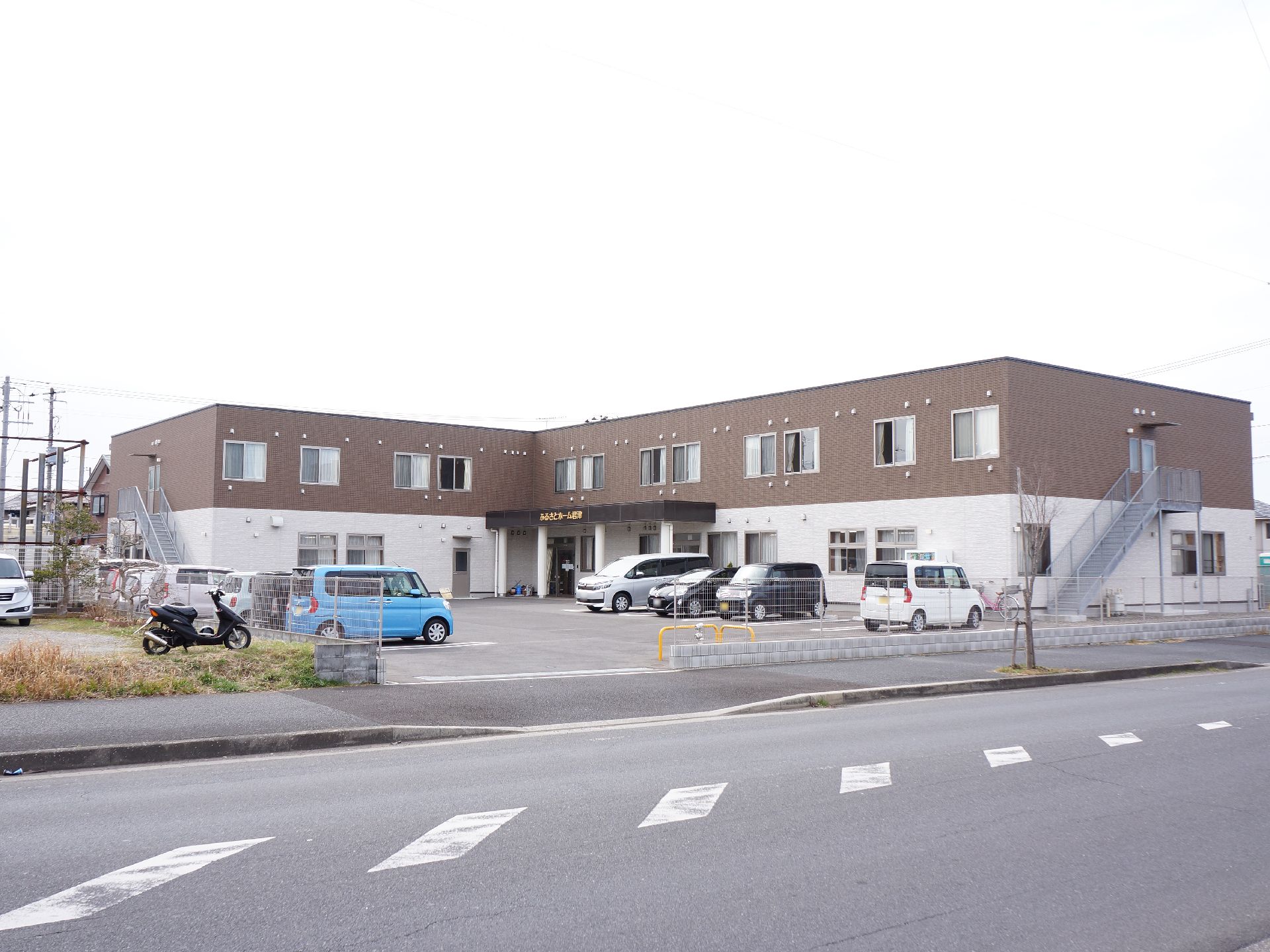 施設は下が白で上が茶色の2階建ての建物。端には非常階段が設置されている。手前に敷地は駐車場になっていて乗用車が数台駐車されている。