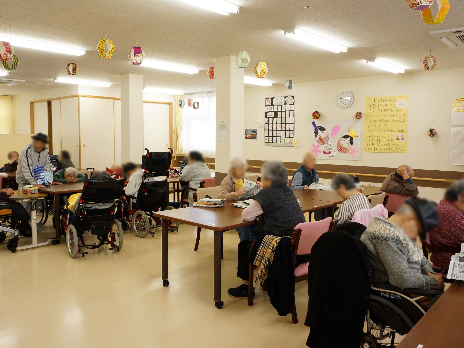 リビングは広く壁には手すりがある。テーブルが多数置かれていてたくさんの入居者が談話している様子が写されている。
