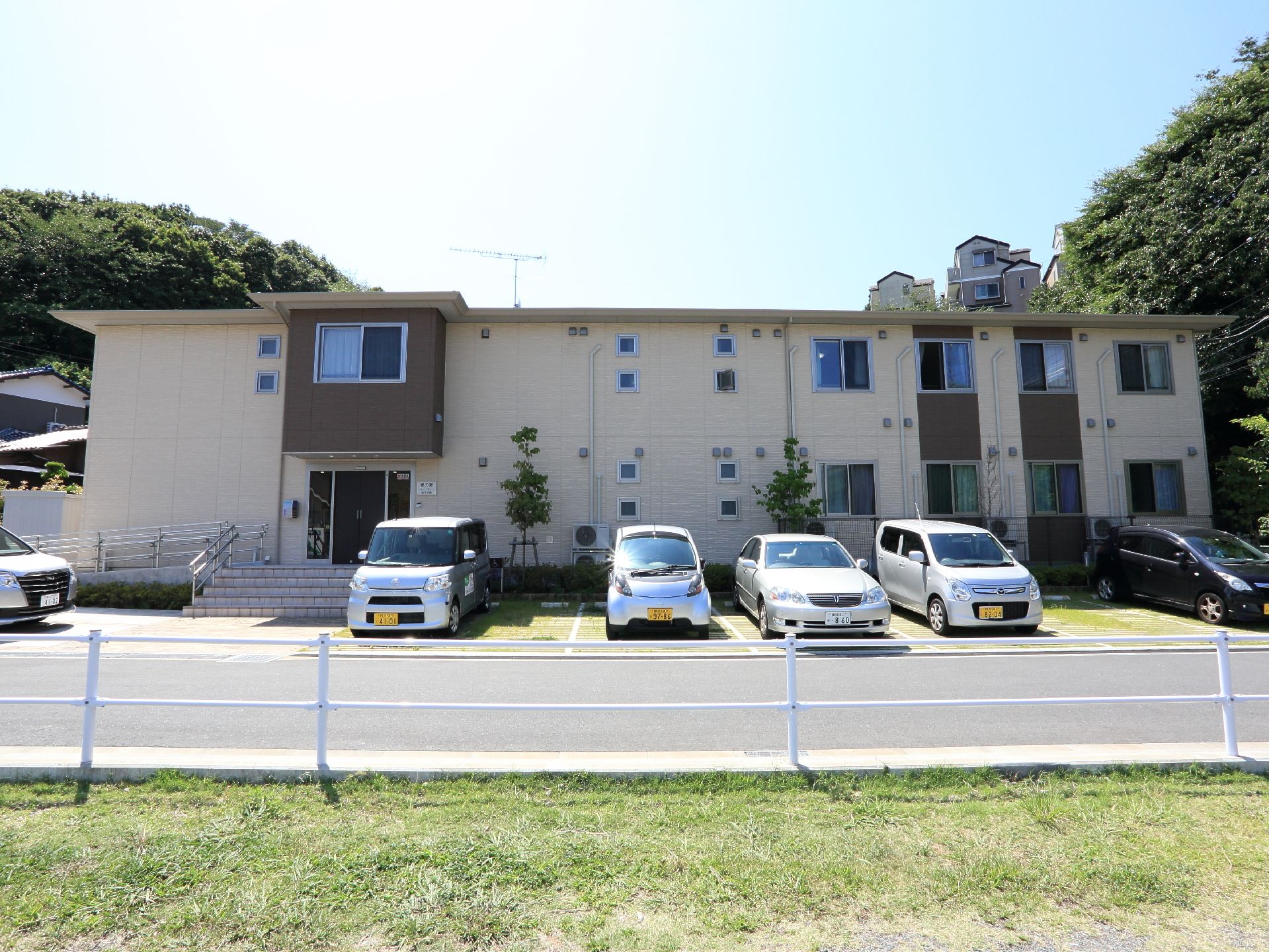 駐車場が2階建の施設の前に設けられている。手すりを備えたスロープと階段がエントランス部分に設置されている。