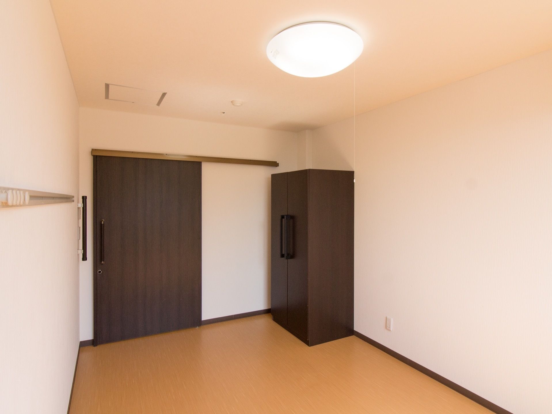 お一人様用の居室である。濃いブラウン色の引き戸と同じ色のロッカータンスが設けられている。ピクチャーレールと電灯が付いている。