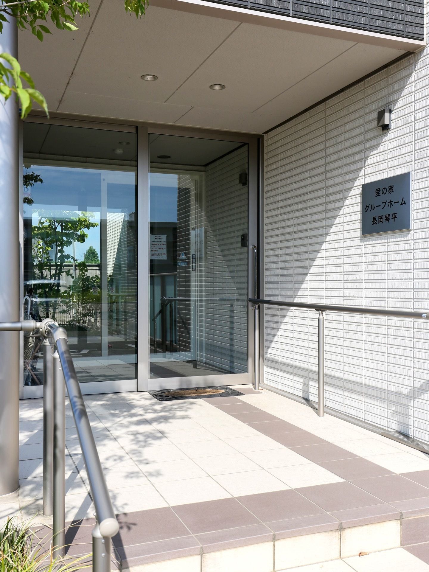 明るい色合いのエントランスである。玄関の扉口にマットが置かれている。両わきには、手すりも設置されている。