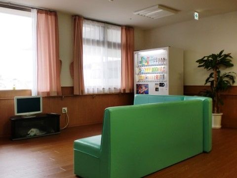 大きな窓にカーテンの掛かった明るいスペースに、コーナーソファとテレビが置かれている。壁際には飲料の自動販売機がある。