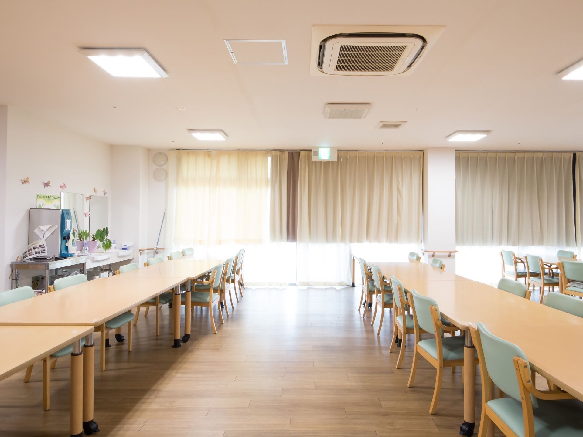 広々とした食堂の奥には、グレーのカーテンの掛けられた窓が取りつけられている。中央には、複数のテーブルや椅子が並べられている。