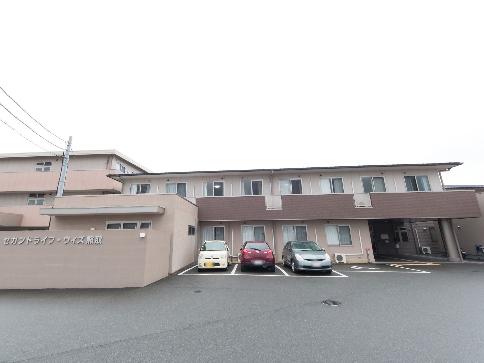 施設は白い色味の外壁で、2階建ての建物となっている。正面の広い駐車場には、3台の車が停められている。