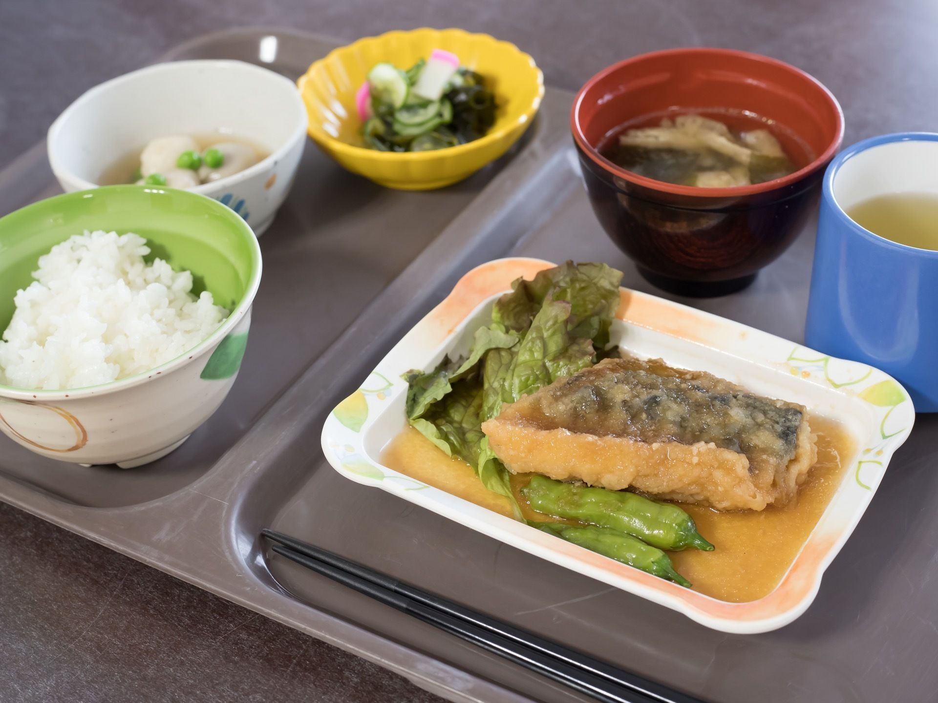 グレーのトレーにはメインの魚料理を中心に、ご飯やみそ汁などが並べられている。黒色のお箸とお茶も添えられている。