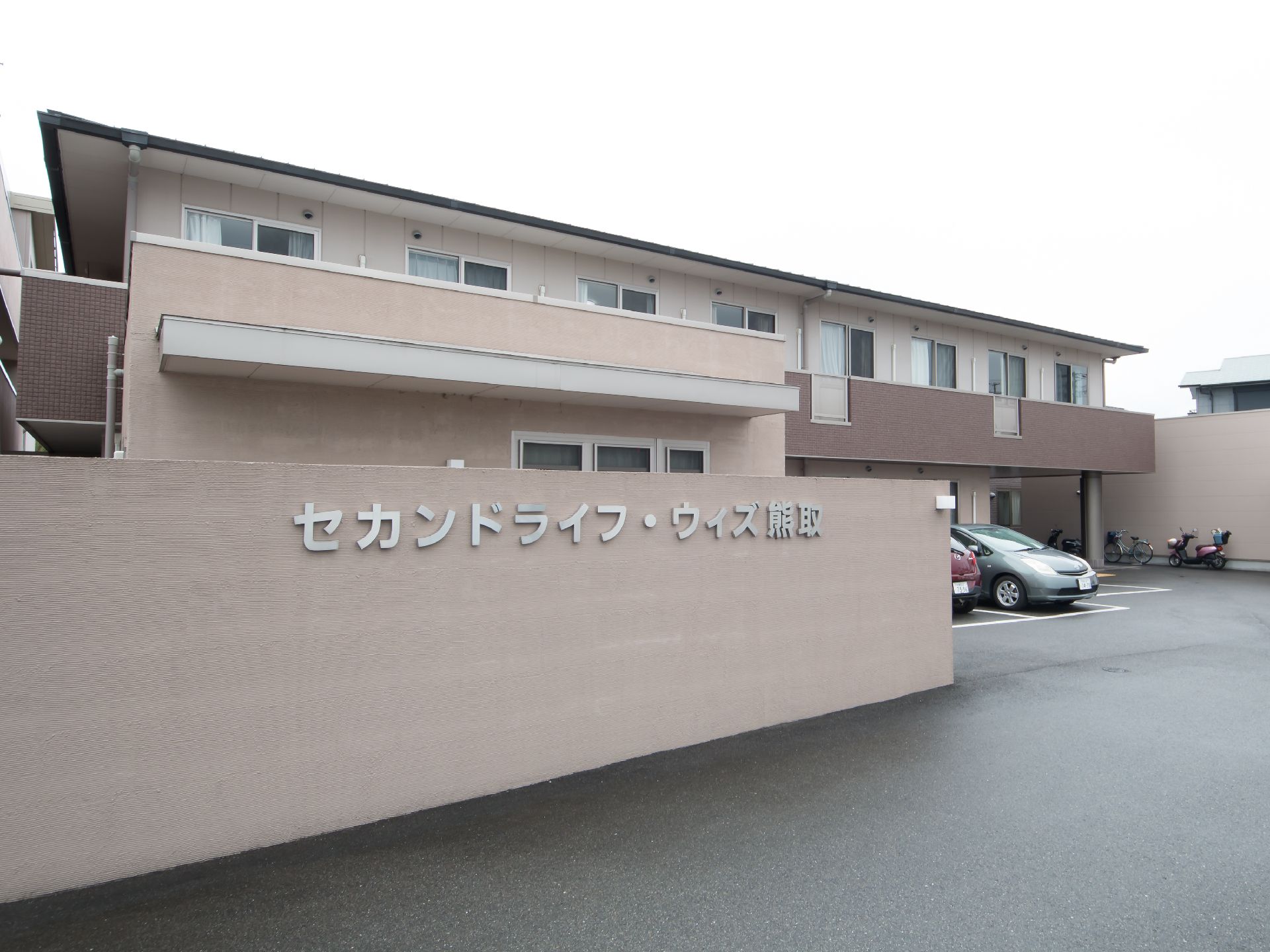 大きく施設名が示された壁があり、駐車場と建物を囲んだ２階建ての施設