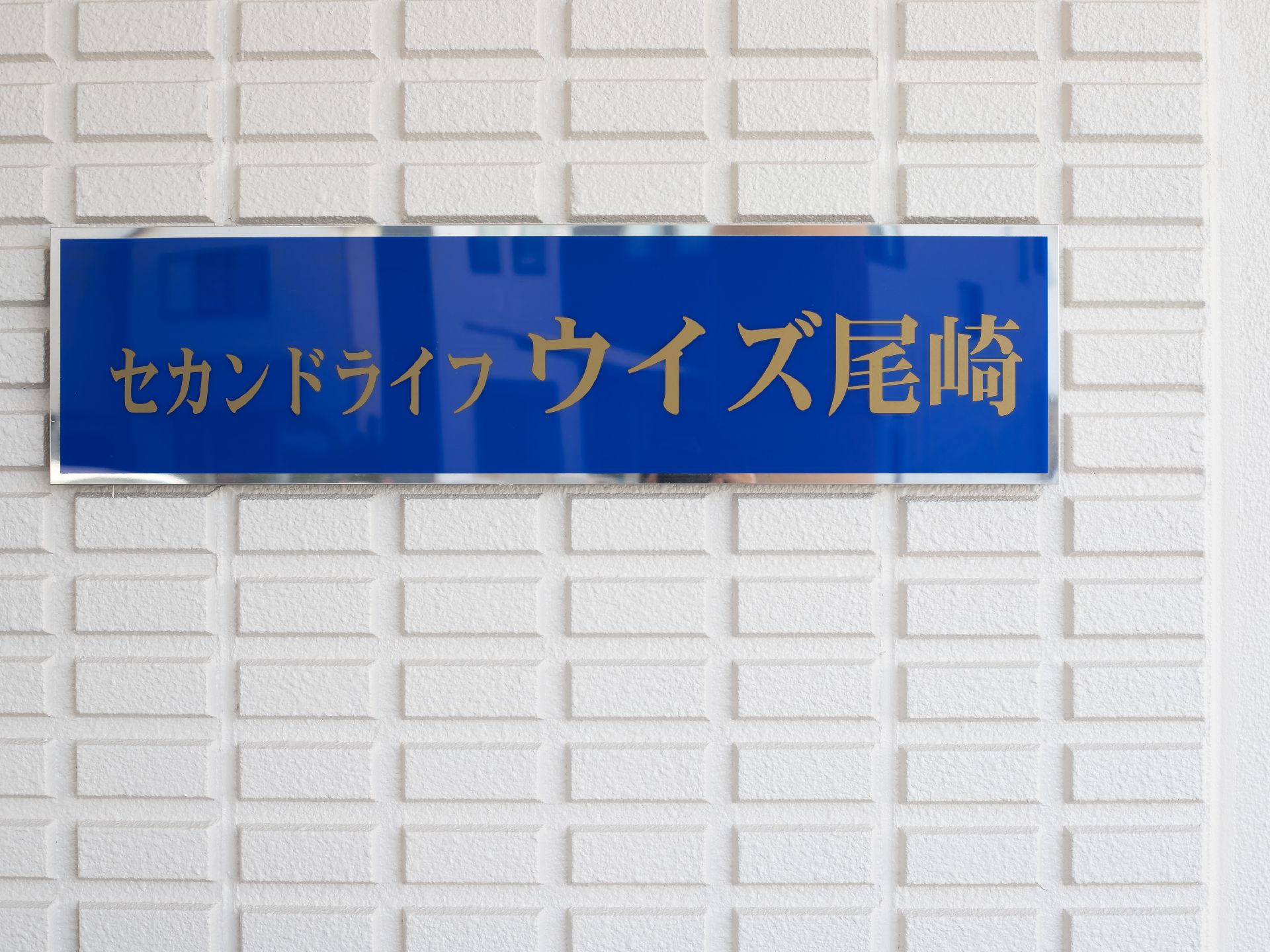 白い壁に青い看板が設置されている。ベージュ色の文字で施設名が書かれていて、看板の周りにはシルバーの縁取りがある。