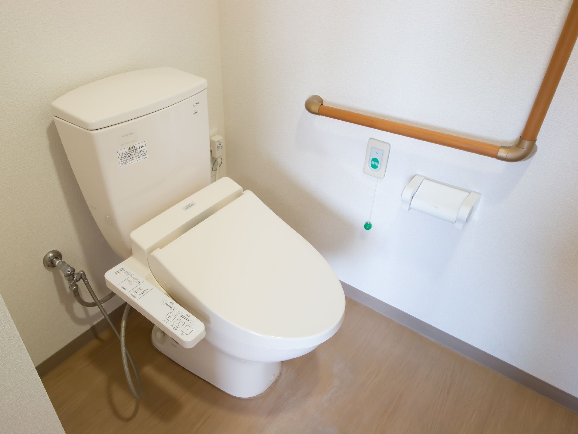 フローリングの床に白いトイレが設置されている。壁に木製の手すりとペーパーホルダー、グリーンの緊急コールが取りつけられている。
