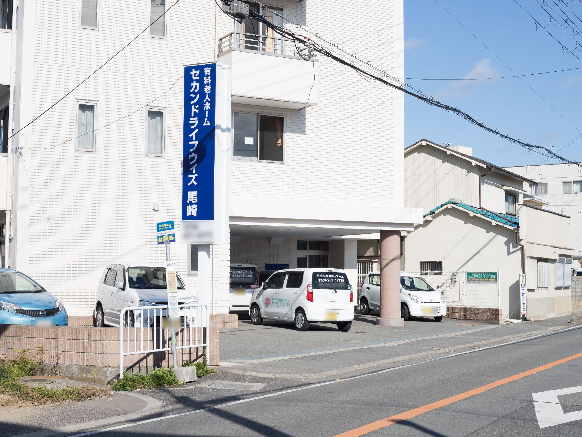 白い外壁の建物で、青い大きな看板が設置されている。建物の前には道路があり、すぐ横にバス停があるのが見える。