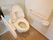 蓋が開いた白いトイレが設置されていて、横に温水洗浄便座の操作スイッチがある。壁には白い手すりが取りつけられている。